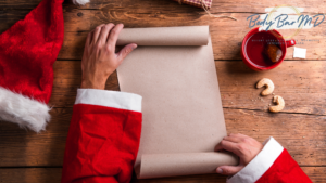 Santa’s hands unrolling a blank parchment on a wooden table surrounded by cookies, tea, and a Santa hat.