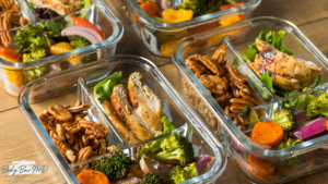 Glass meal prep containers filled with grilled chicken, roasted vegetables, and pecans, arranged on a wooden surface.