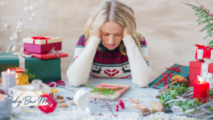 A stressed woman wearing a festive sweater sits at a holiday-decorated table, surrounded by gifts and decorations.