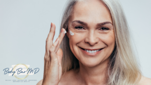 Smiling mature woman with long silver hair applying moisturizer under her eye, promoting skincare and healthy aging