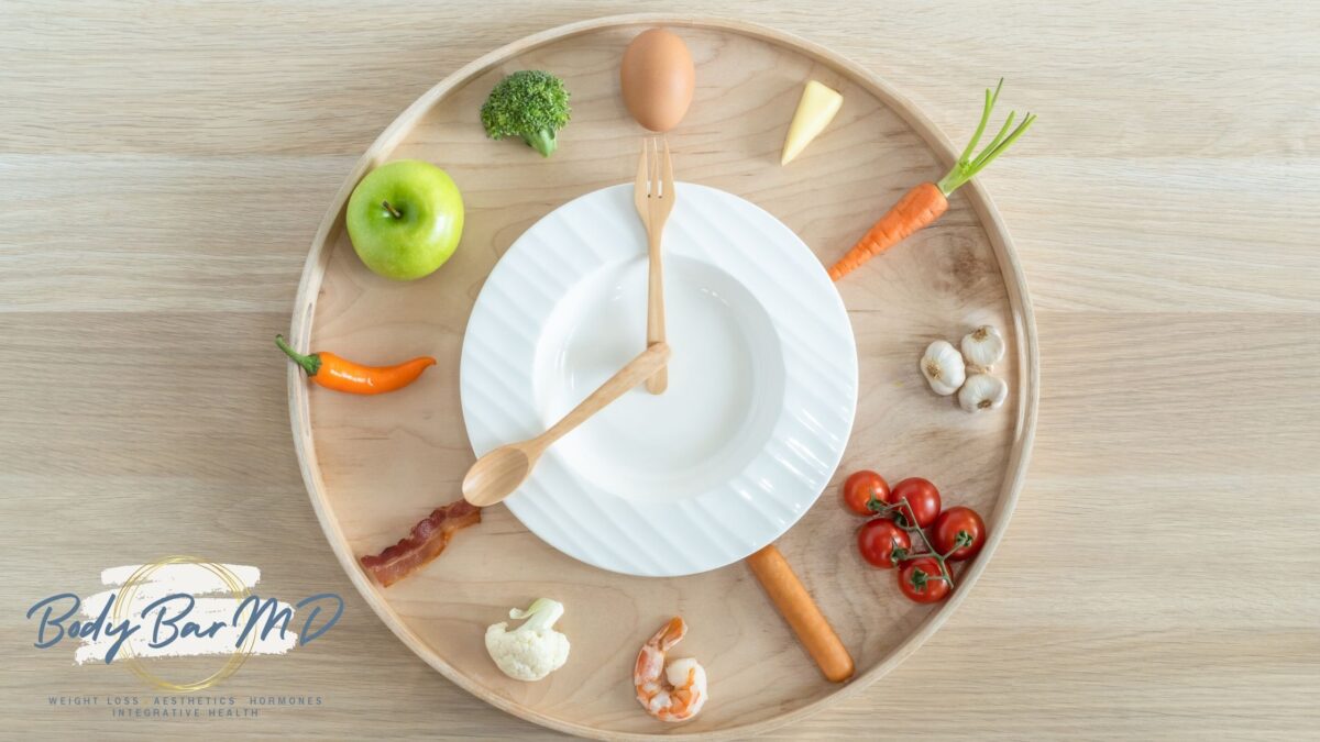 A plate and wooden utensils arranged as a clock, surrounded by fresh vegetables, fruits, and protein-rich foods.