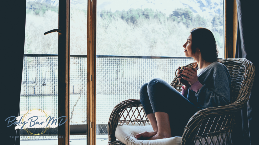 Woman sitting in a wicker chair by a window, holding a mug, enjoying a serene winter morning.