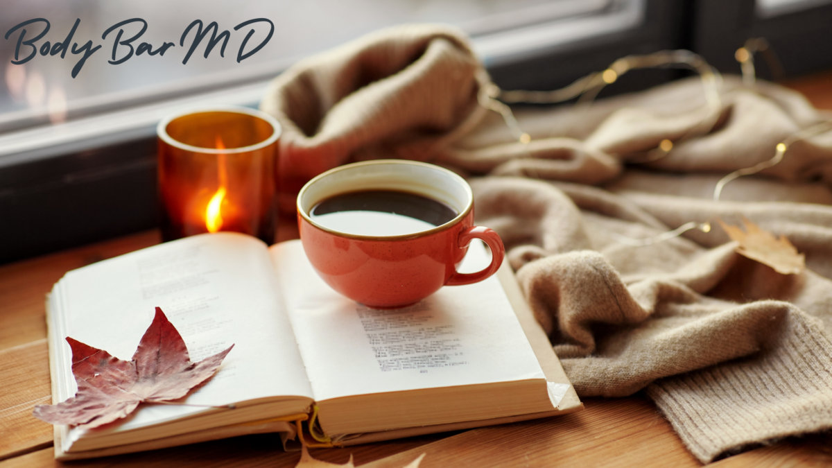 A cozy autumn scene featuring an open book with a dried maple leaf on its pages, a warm cup of coffee in a red mug, and a softly lit candle in the background. A soft beige sweater and string lights add warmth, with Body Bar MD branding subtly visible. The scene evokes a calm, inviting atmosphere, ideal for relaxation and wellness