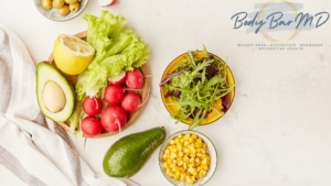 Fresh and vibrant healthy foods including lettuce, radishes, avocado, lemon, corn, and mixed greens arranged on a light background with the Body Bar MD logo, representing the focus on nutritious, whole foods for wellness