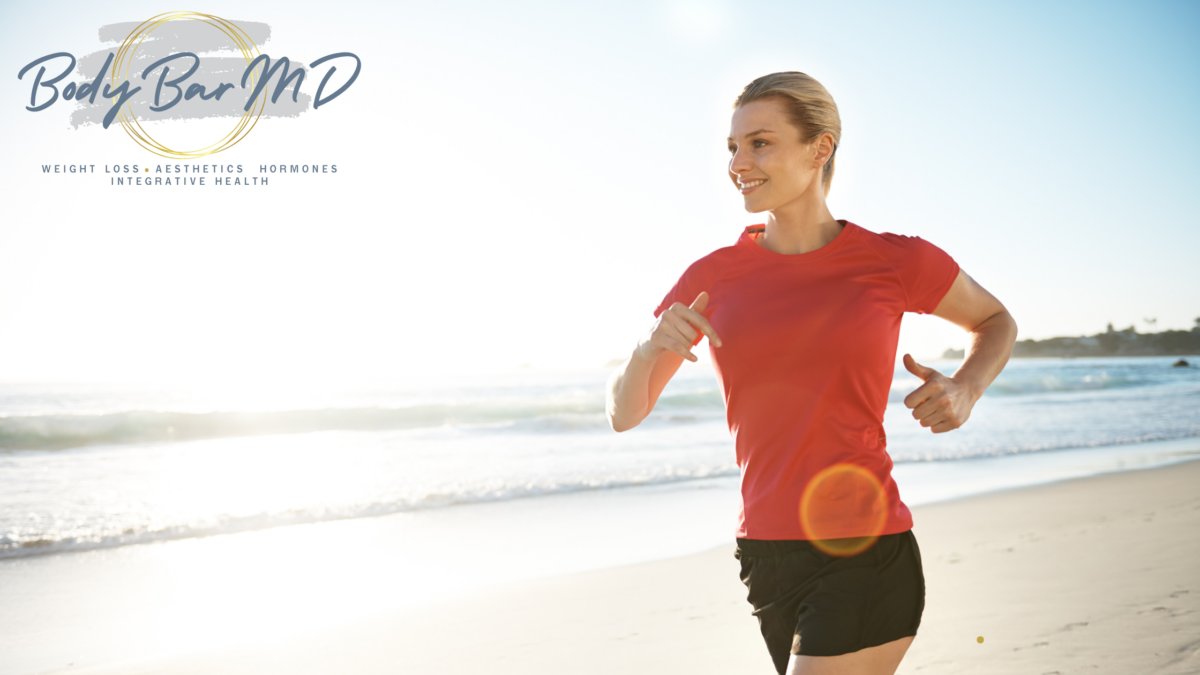 Woman jogging on a beach in a red shirt, smiling with energy, with the Body Bar MD logo in the corner. Represents vitality, fitness, and well-being.