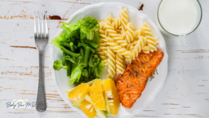 A balanced meal of salmon, pasta, greens, and orange slices on a white plate with a glass of milk