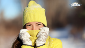 A woman bundled up in a bright yellow hat and scarf, smiling outdoors on a snowy winter day.