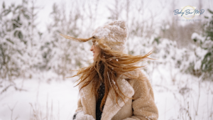 A woman dressed in a cozy beige coat and hat smiles as her hair blows in the snowy winter landscape.