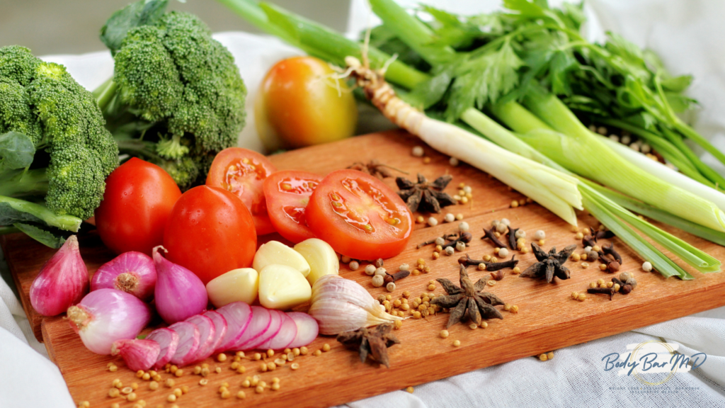 A variety of fresh vegetables and herbs, including broccoli, tomatoes, onions, and spices, on a wooden cutting board.