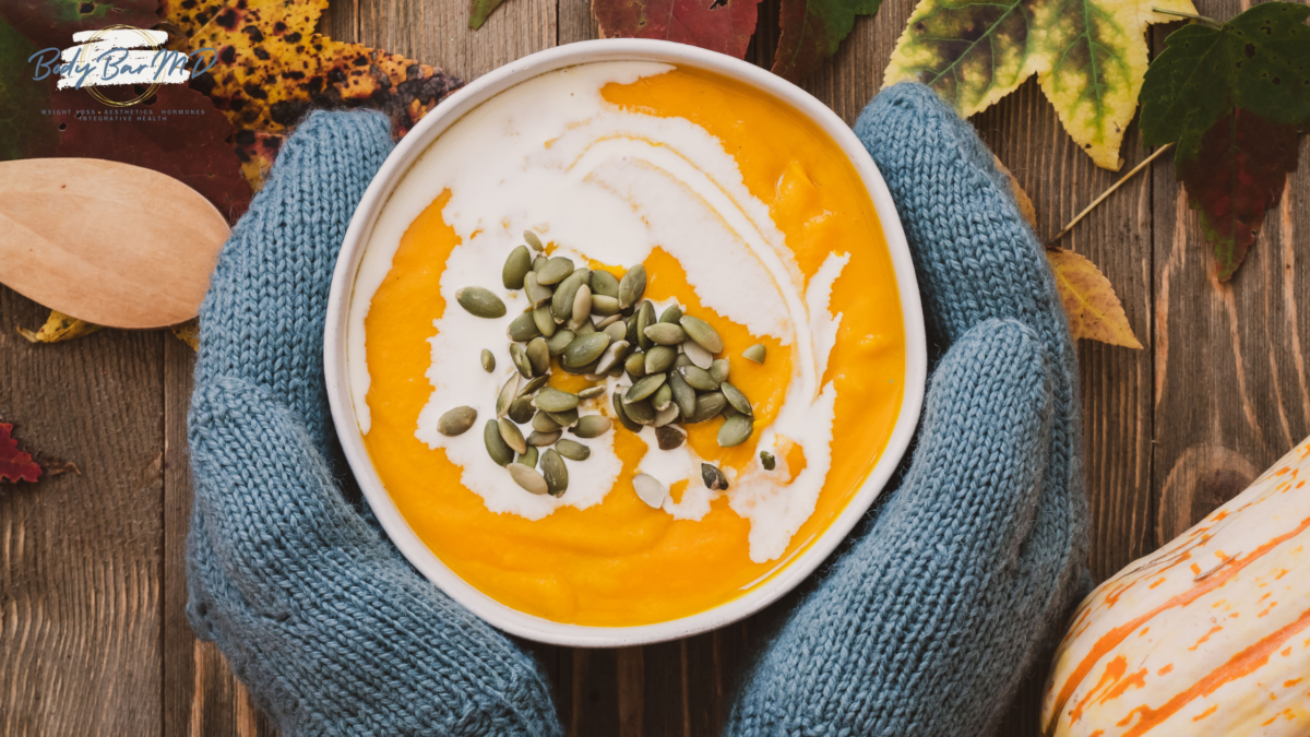 A pair of blue-gloved hands holding a bowl of pumpkin soup topped with cream and pumpkin seeds.