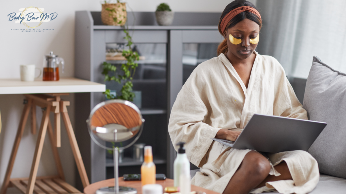 A woman in a robe with gold eye patches works on a laptop during a self-care session at home.