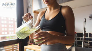 A woman in a sports bra pours a green smoothie into a glass, standing in a bright kitchen.