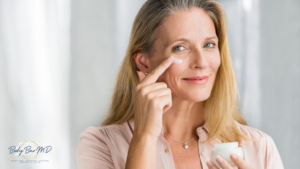 A smiling middle-aged woman applying moisturizer under her eye, representing youthful, glowing skin.