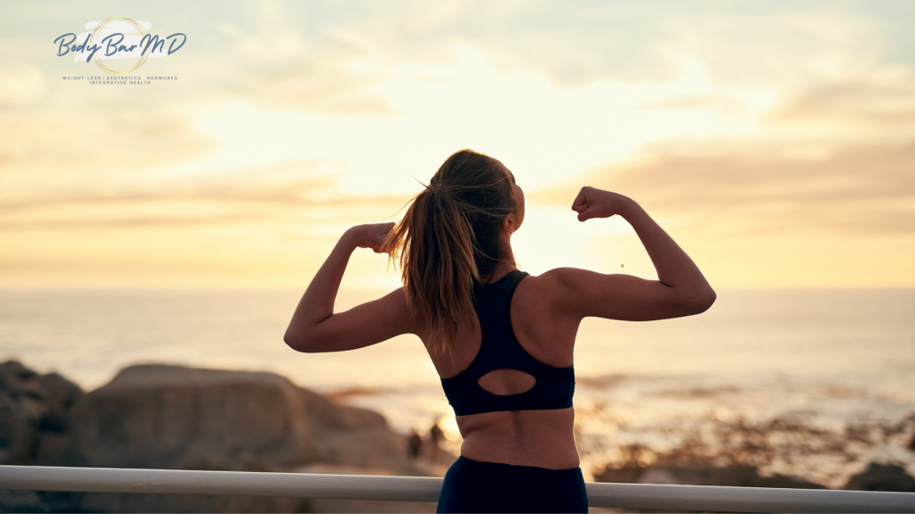 A woman flexing her arms in a strong pose, overlooking a scenic sunset by the ocean.