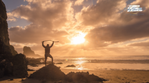 A silhouette of a person raising their arms triumphantly on a rocky beach at sunset, symbolizing empowerment and achievement. Energized after a B12 Shot