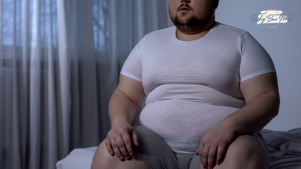 Overweight man sitting on a bed, wearing a white shirt and gray shorts, reflecting a sedentary lifestyle.