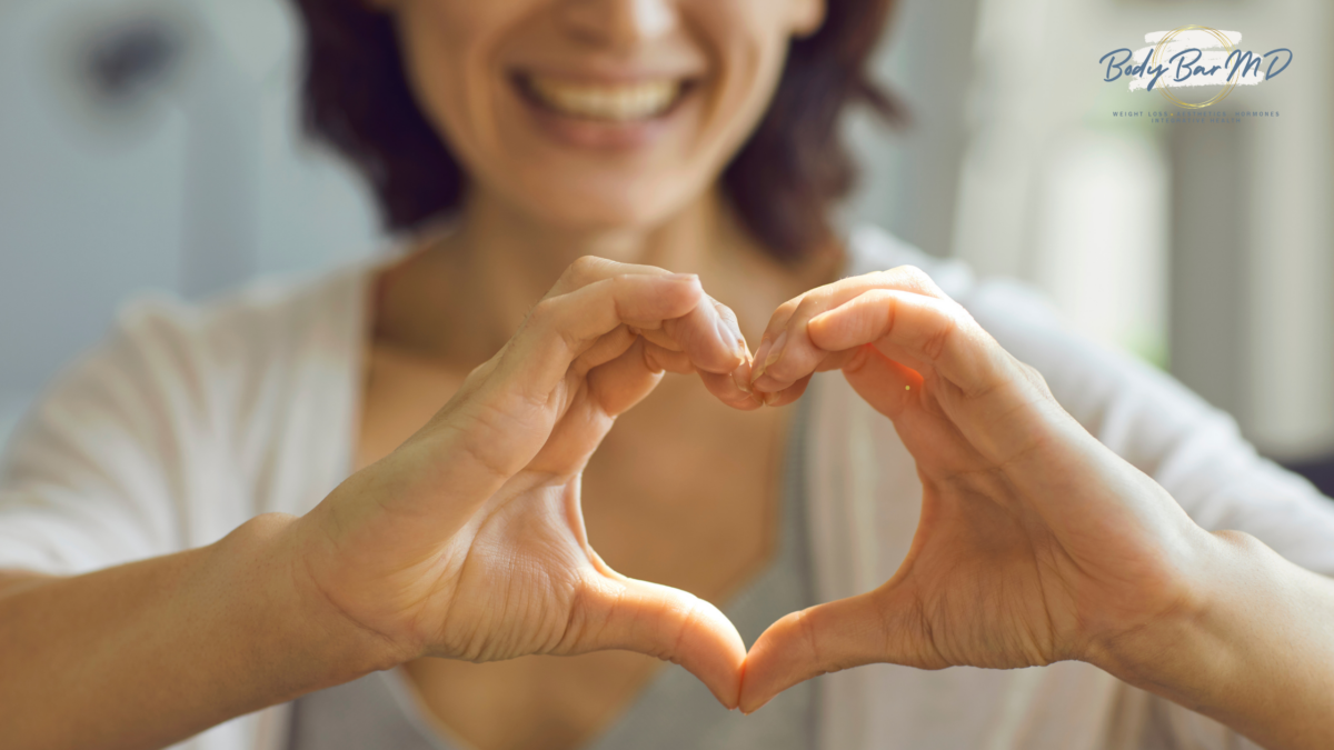 Smiling woman forming a heart shape with her hands, symbolizing heart health and self-love. The Body Bar MD logo is visible in the corner.