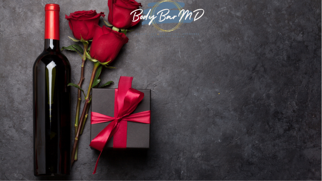 A romantic flat lay featuring a bottle of wine, three red roses, and a black gift box tied with a red satin ribbon, with the Body Bar MD logo at the top.