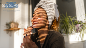 Smiling woman applying skincare with a towel wrapped around her hair, sunlight streaming through blinds, featuring the Body Bar MD logo.