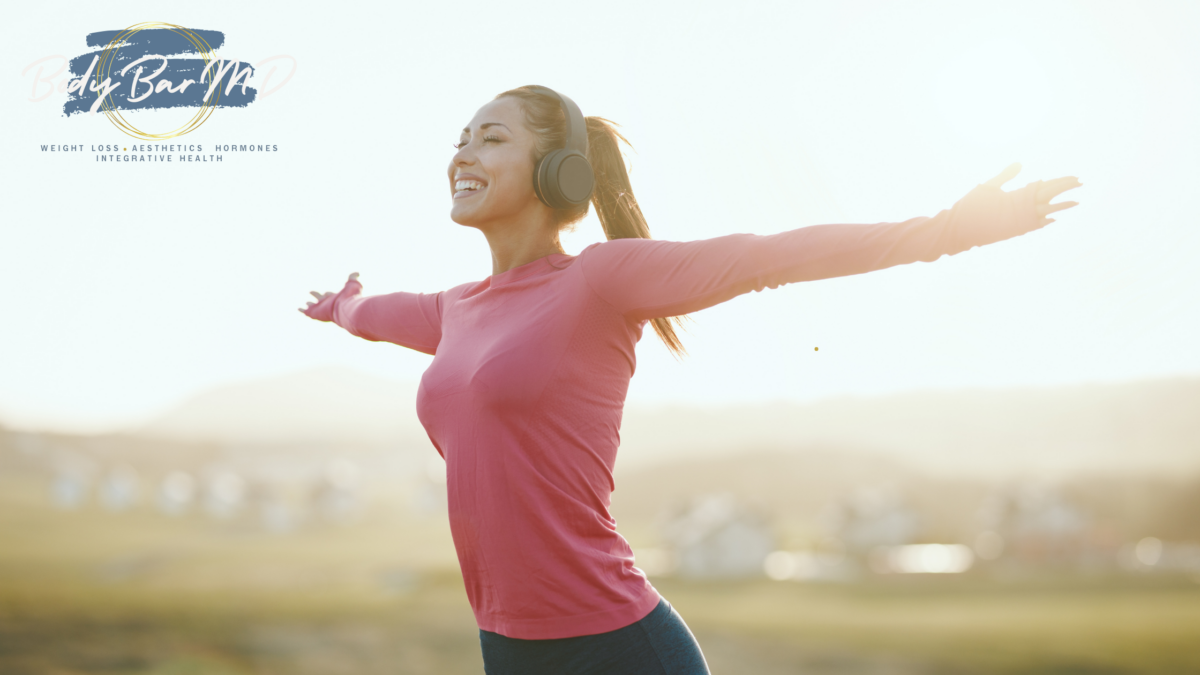 Smiling woman in a pink workout top with arms outstretched, enjoying the outdoors and sunshine, wearing headphones. Body Bar MD logo in the corner.