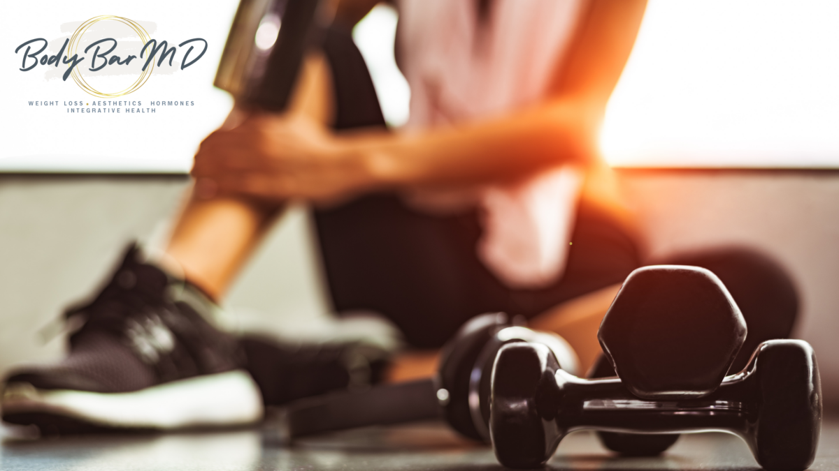 Close-up of dumbbells on a gym floor with a person resting after a workout, sunlight streaming through the window, and Body Bar MD logo in the corner.