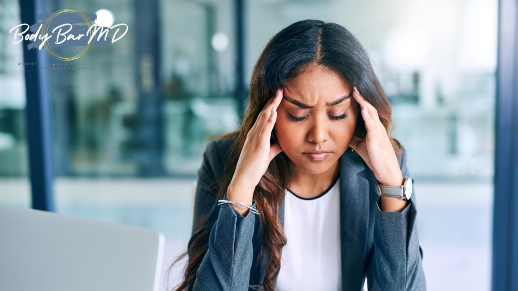 A woman in a business suit sitting at a desk with her hands on her temples, appearing stressed and overwhelmed.