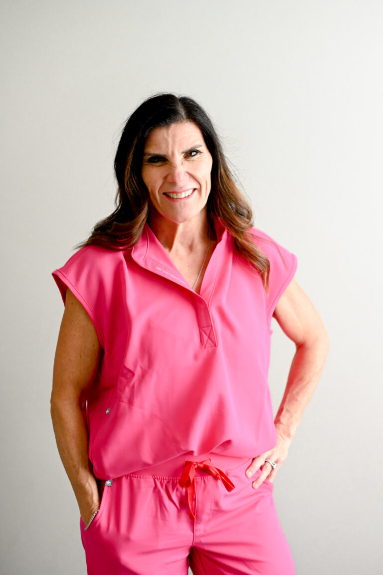 A confident woman wearing a bright pink medical uniform smiles warmly, standing with her hands on her hips, projecting professionalism and approachability.